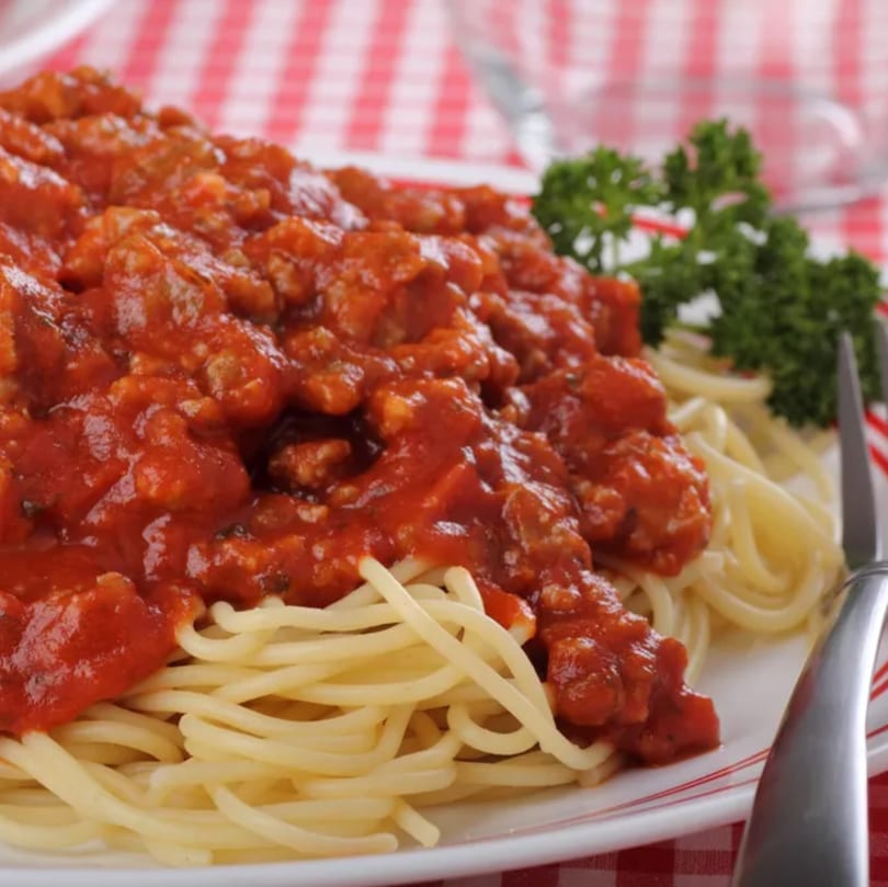 Spaghetti with rich red meat sauce and a sprig of parsley on a white plate with red checkered tablecloth.