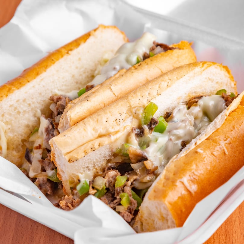 Two cheesy Philly cheesesteak sandwiches with peppers and onions in a white paper-lined container.