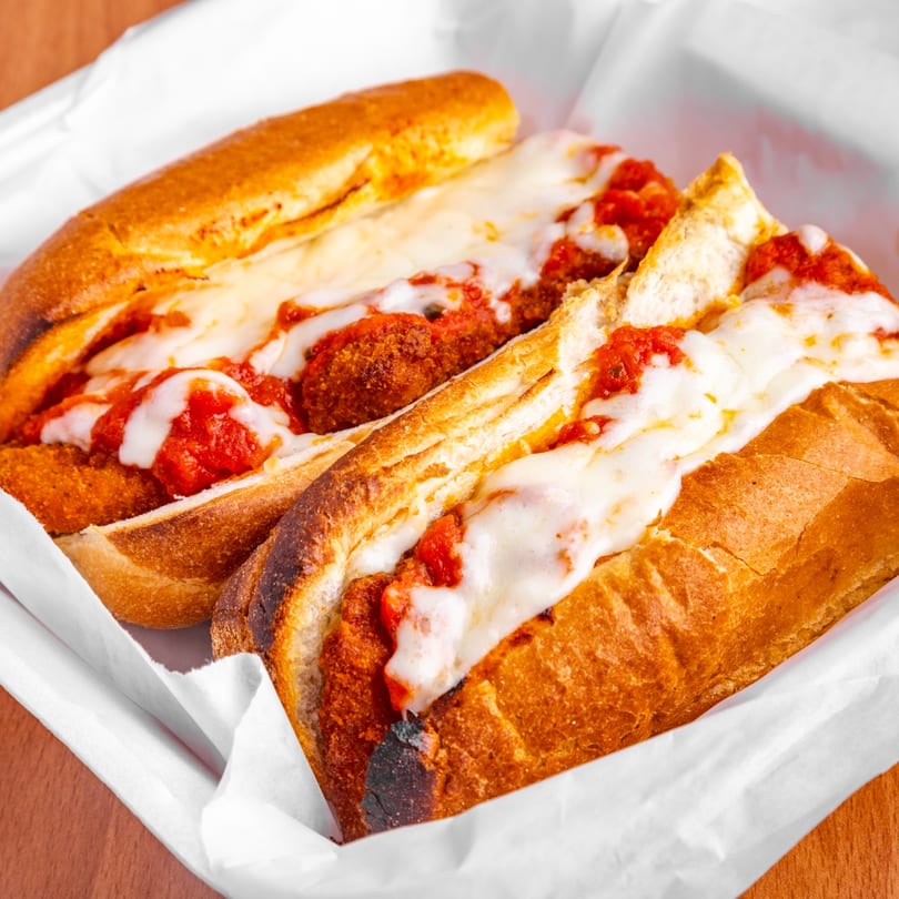 Two meatball parmesan subs with melted cheese and marinara sauce in a white paper-lined basket.